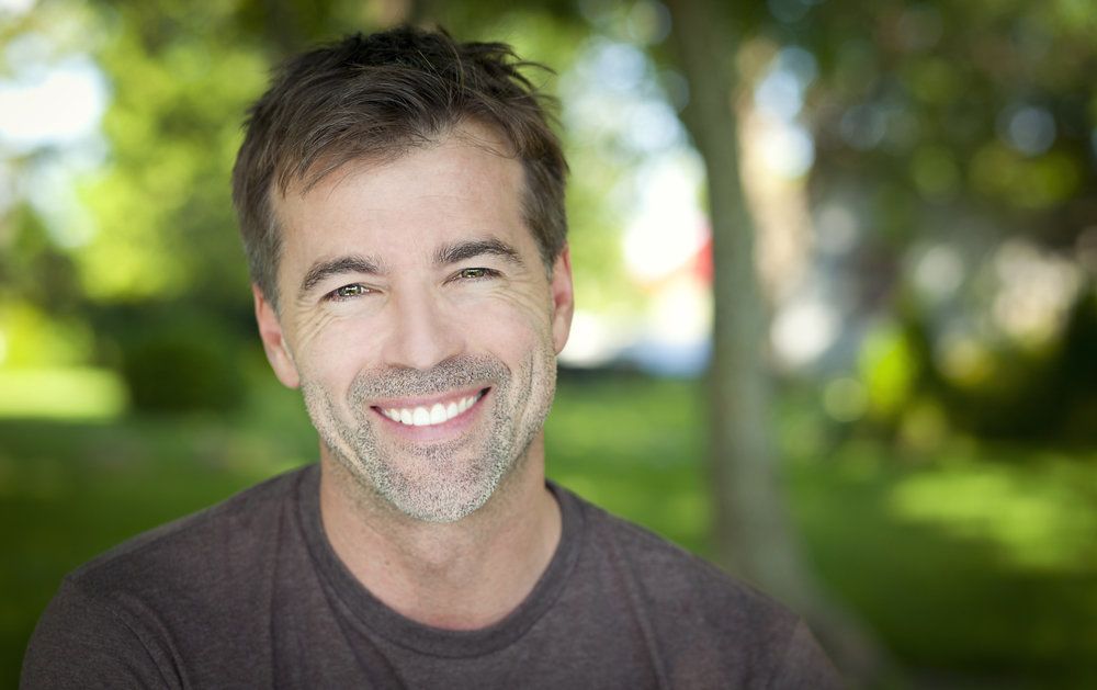 A handsome, middle-aged man with light brown hair and a grayed beard smiles outside.